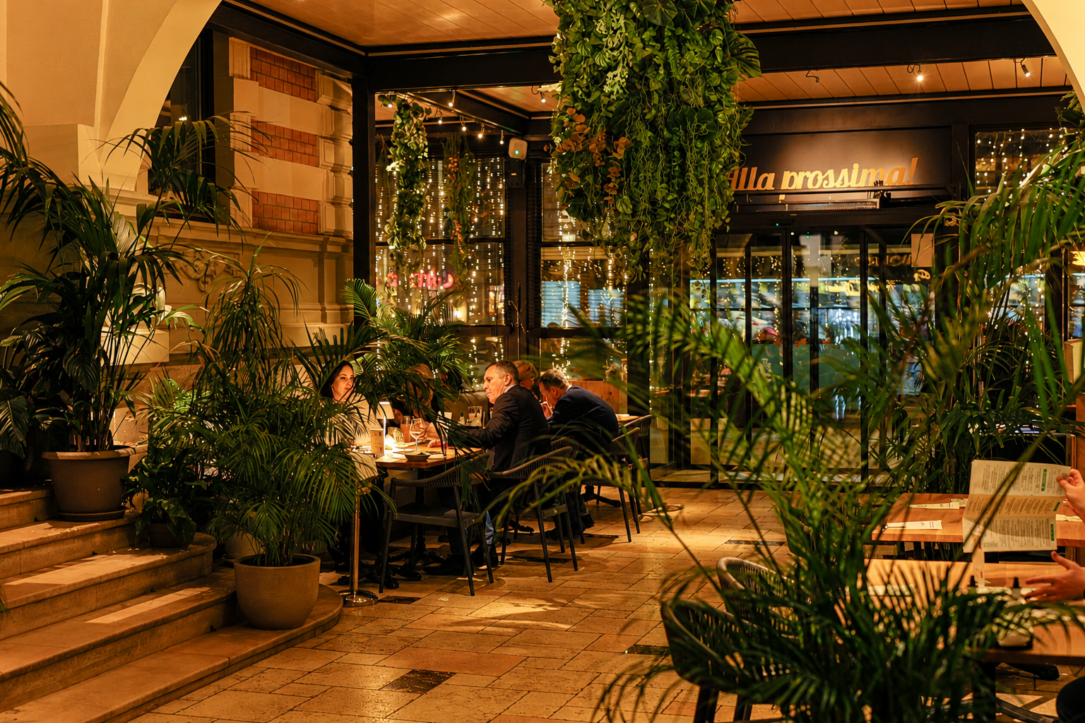 Indoor restaurant dining area with lush potted plants, warm amber lighting, and diners at a table.