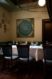 A dining room with white-tablecloth tables set for guests, wooden chairs, wine glasses, and a large blue circular wall art centerpiece.