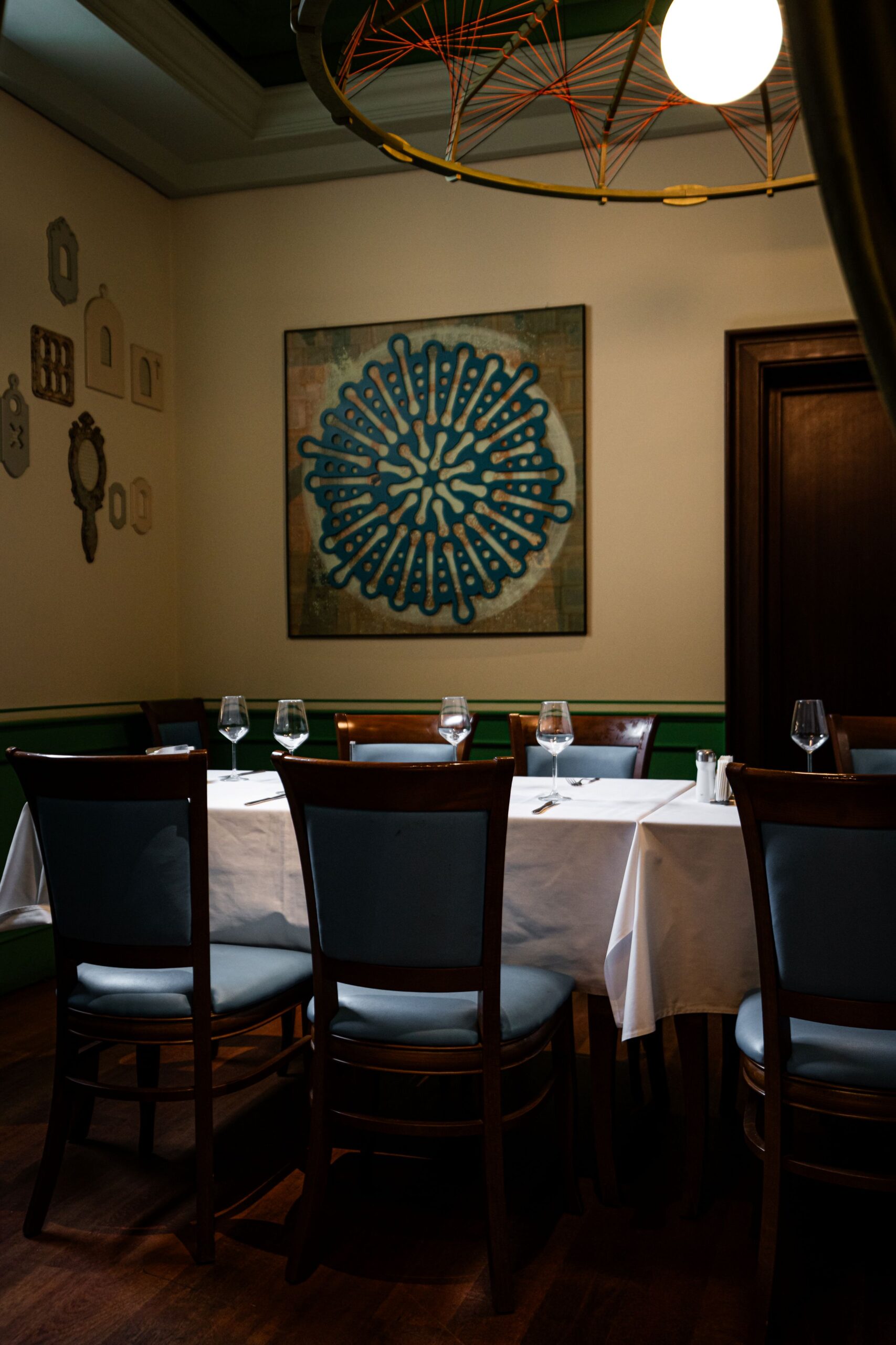 A dining room with white-tablecloth tables set for guests, wooden chairs, wine glasses, and a large blue circular wall art centerpiece.