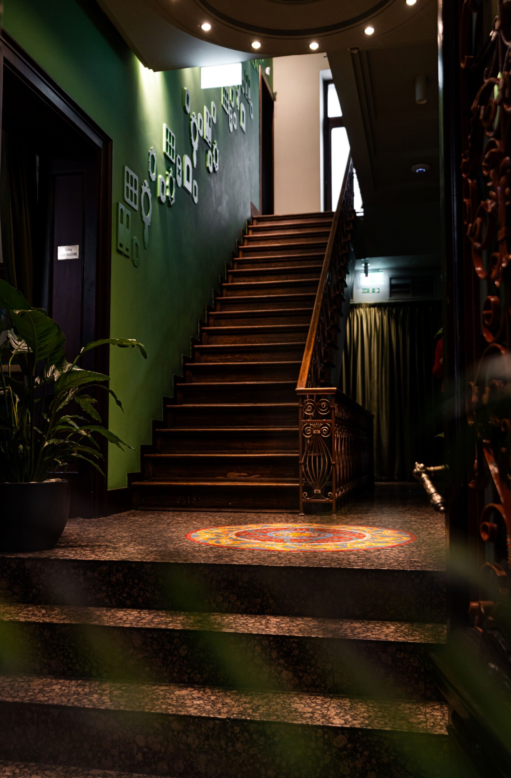 Interior staircase with a green wall, decorative wall shapes, and ornate railing; colorful circular rug at the base; plants on the left and soft overhead lights.