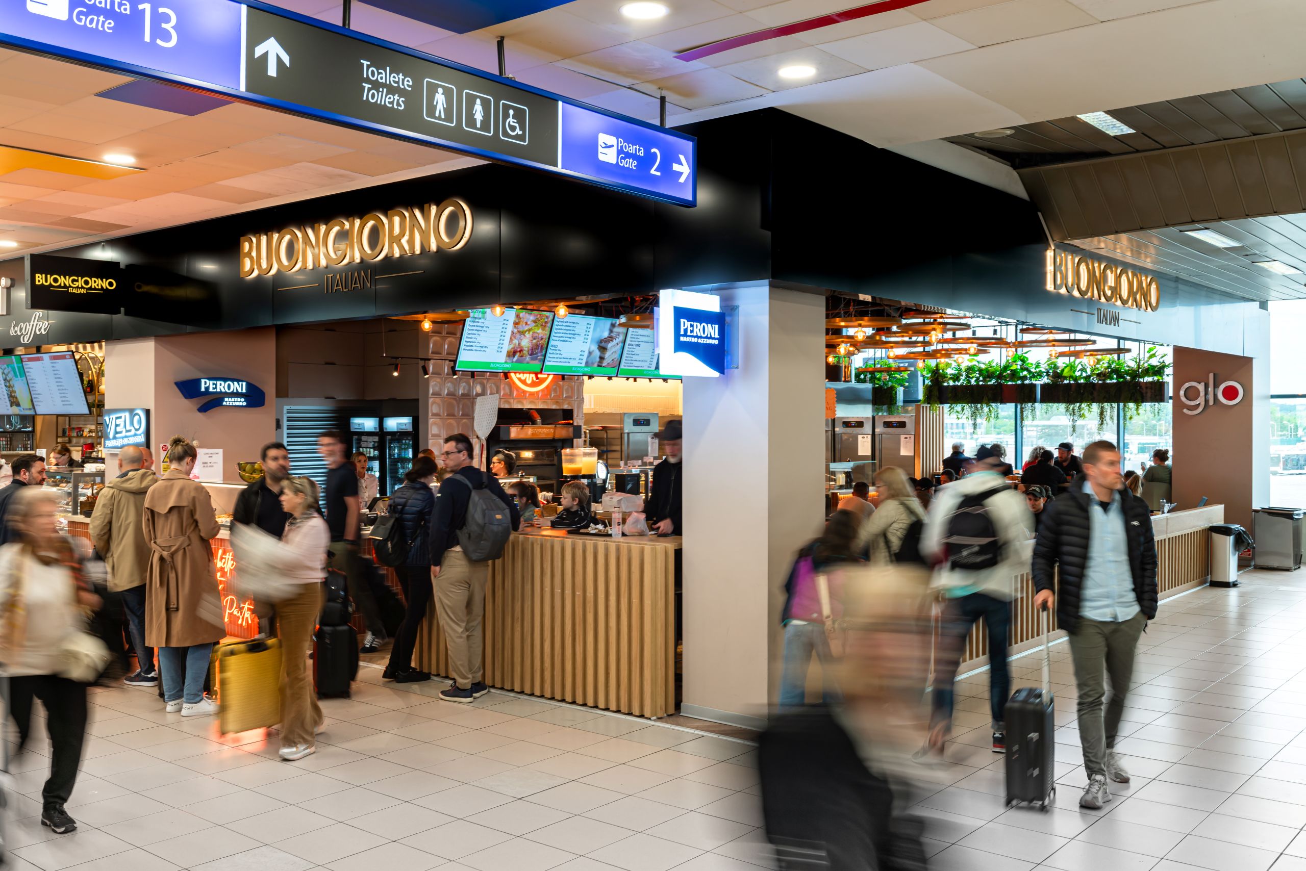 Busy airport terminal with a Buongiorno Italian cafe and digital menu boards amidst travelers with rolling luggage and lines at the counter