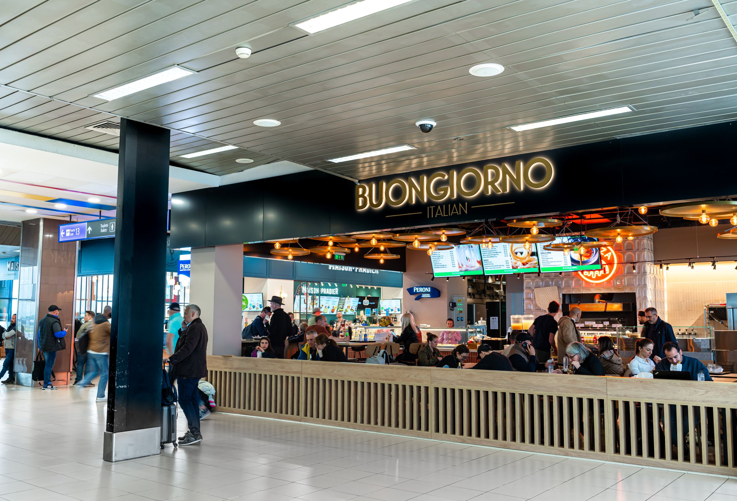Busy airport-style food court with a bright BUONGIORNO ITALIAN sign over a counter and diners seated at tables under warm string lights.