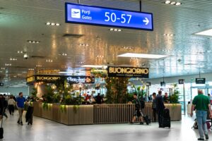 Busy airport concourse with a blue 50–57 gates sign and multiple dining signs, including Buongiorno Italian, amid travelers and plants.