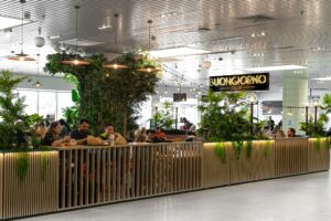 Airport dining area with long wooden booths, lush plants, warm pendant lights, and a Buongiorno Italian sign.