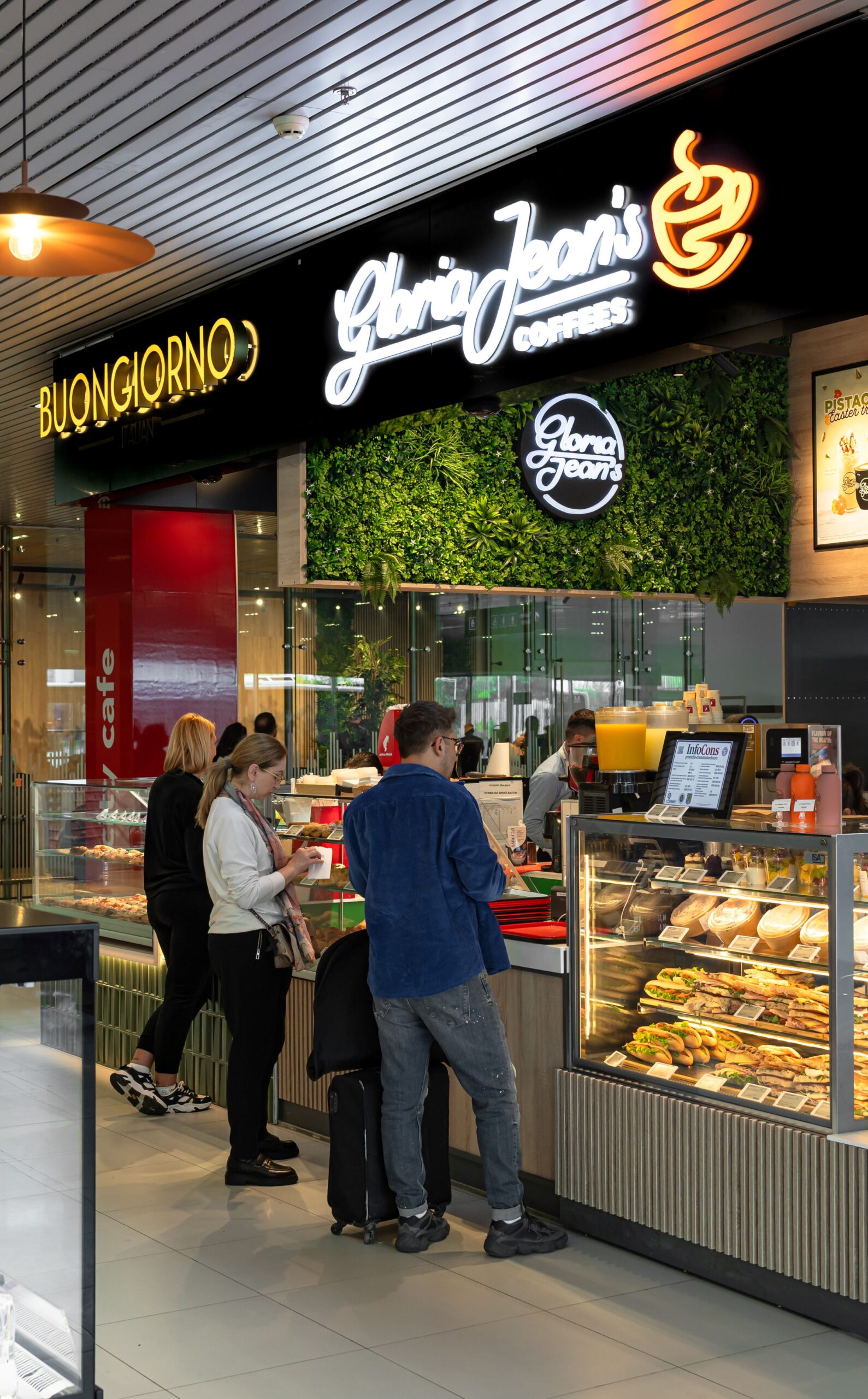 People in a queue at a Gloria Jean's Coffees counter with pastries and drinks on display behind a glass case.