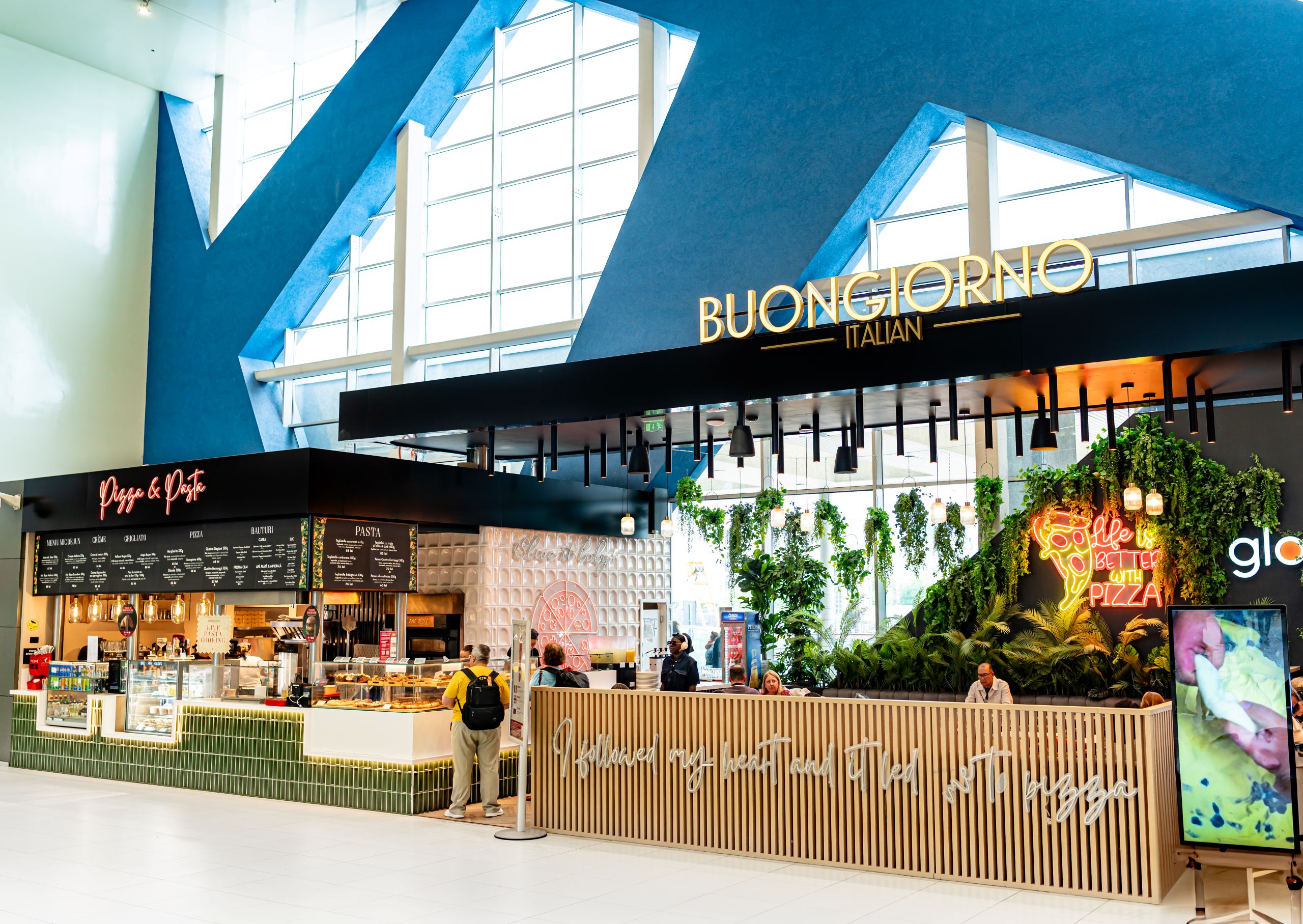 Indoor food court with 'Buongiorno Italian' sign above a pizza counter and greenery throughout the space