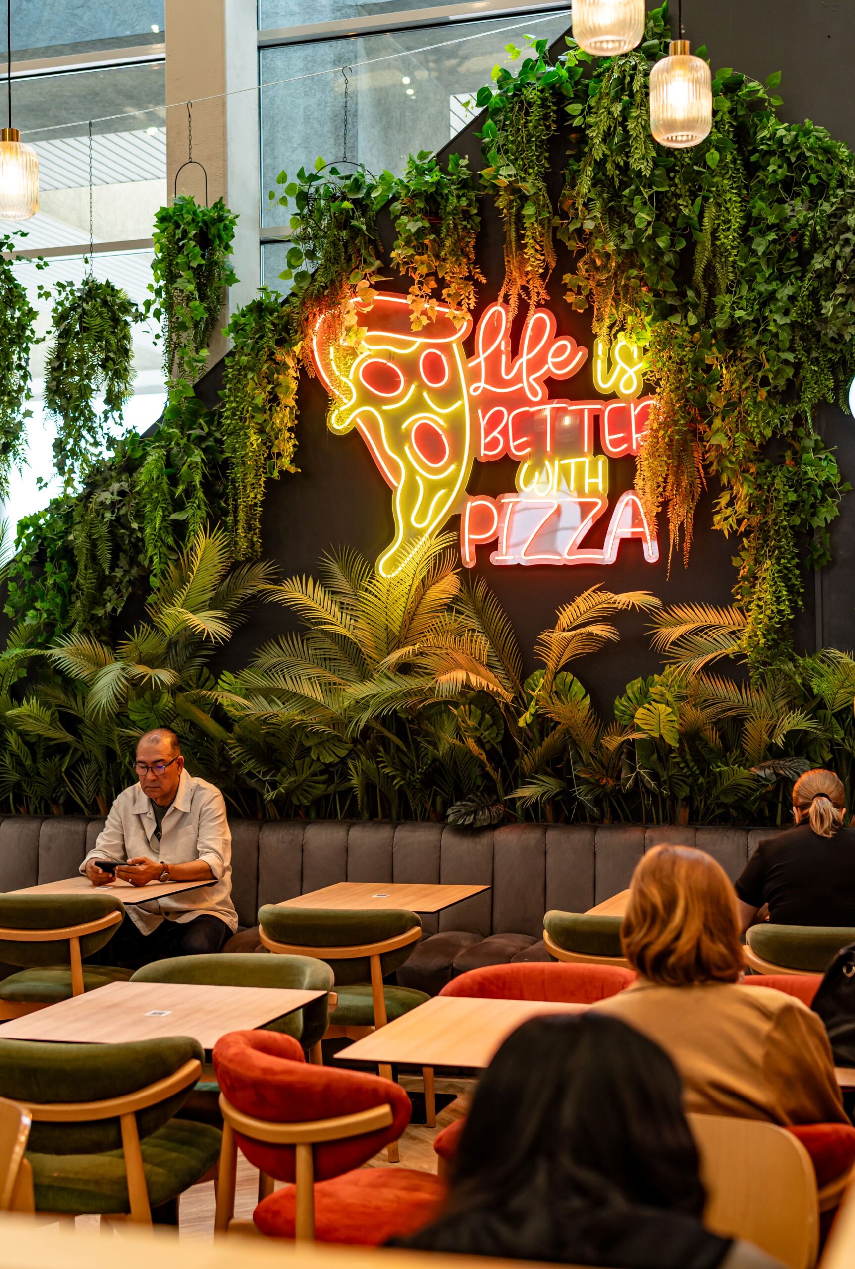 Plant-filled cafe seating area with a bright neon pizza sign on a dark wall; people sit at tables and chat or work nearby.