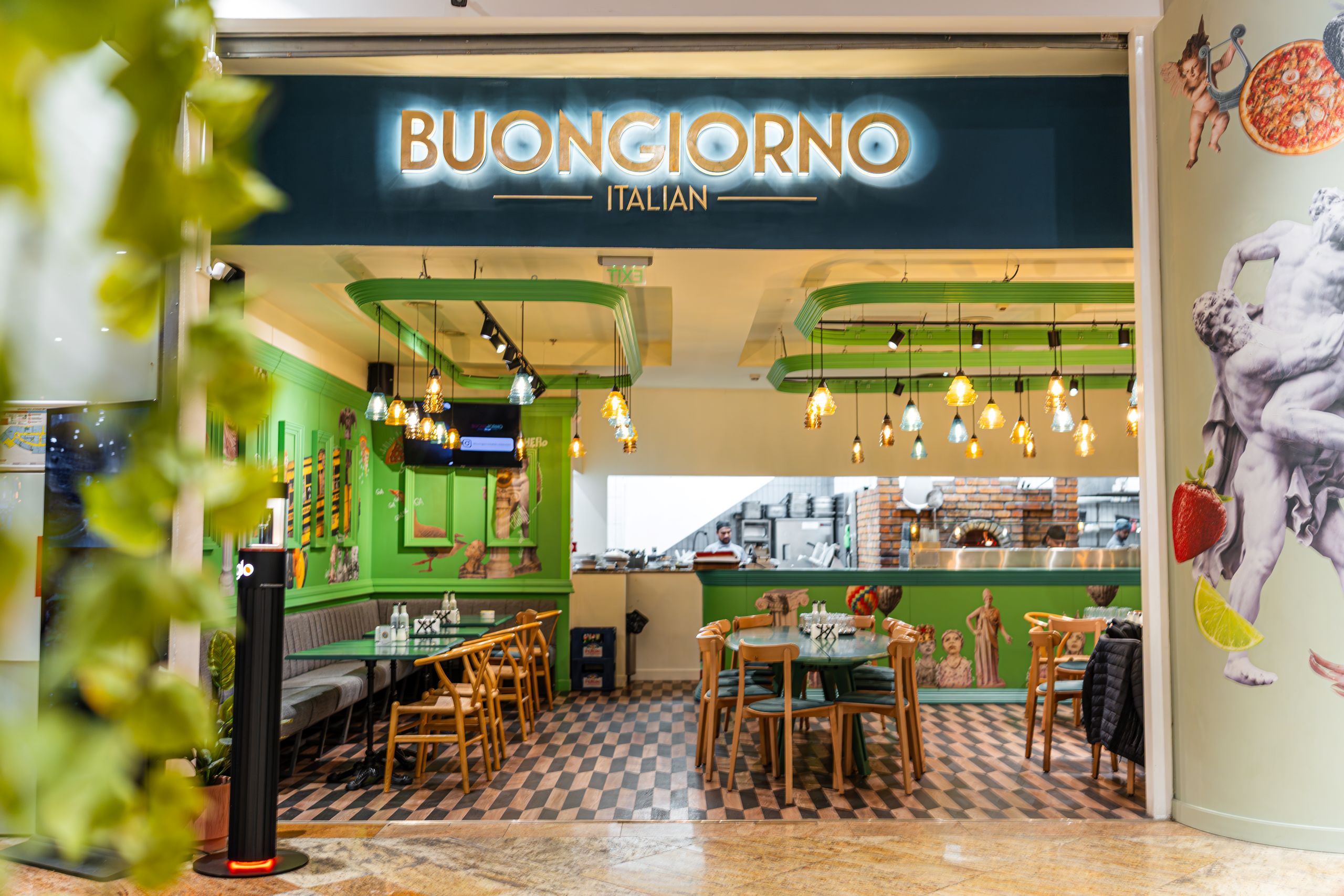 Open Italian restaurant interior with green walls, wooden chairs, and warm pendant lights over a checkered floor.