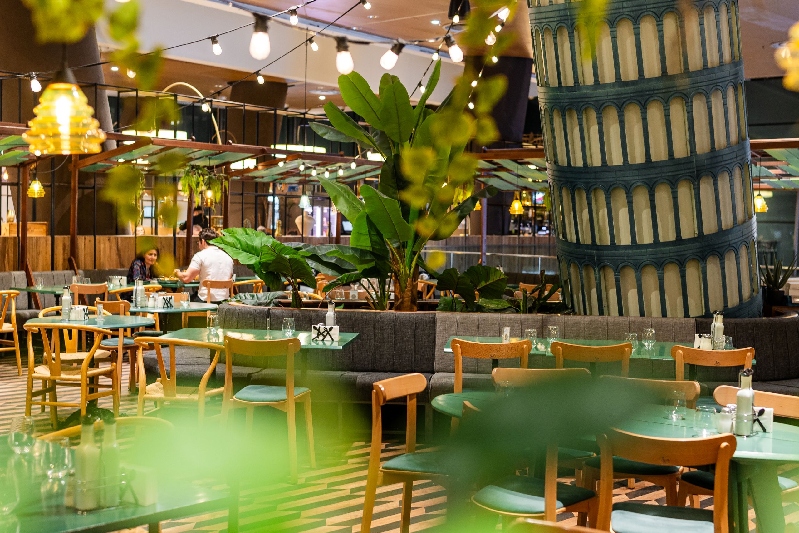 Restaurant dining area with green tables, wooden chairs, and tropical plants; string lights above and a large blue-tiled cylindrical pillar on the right.