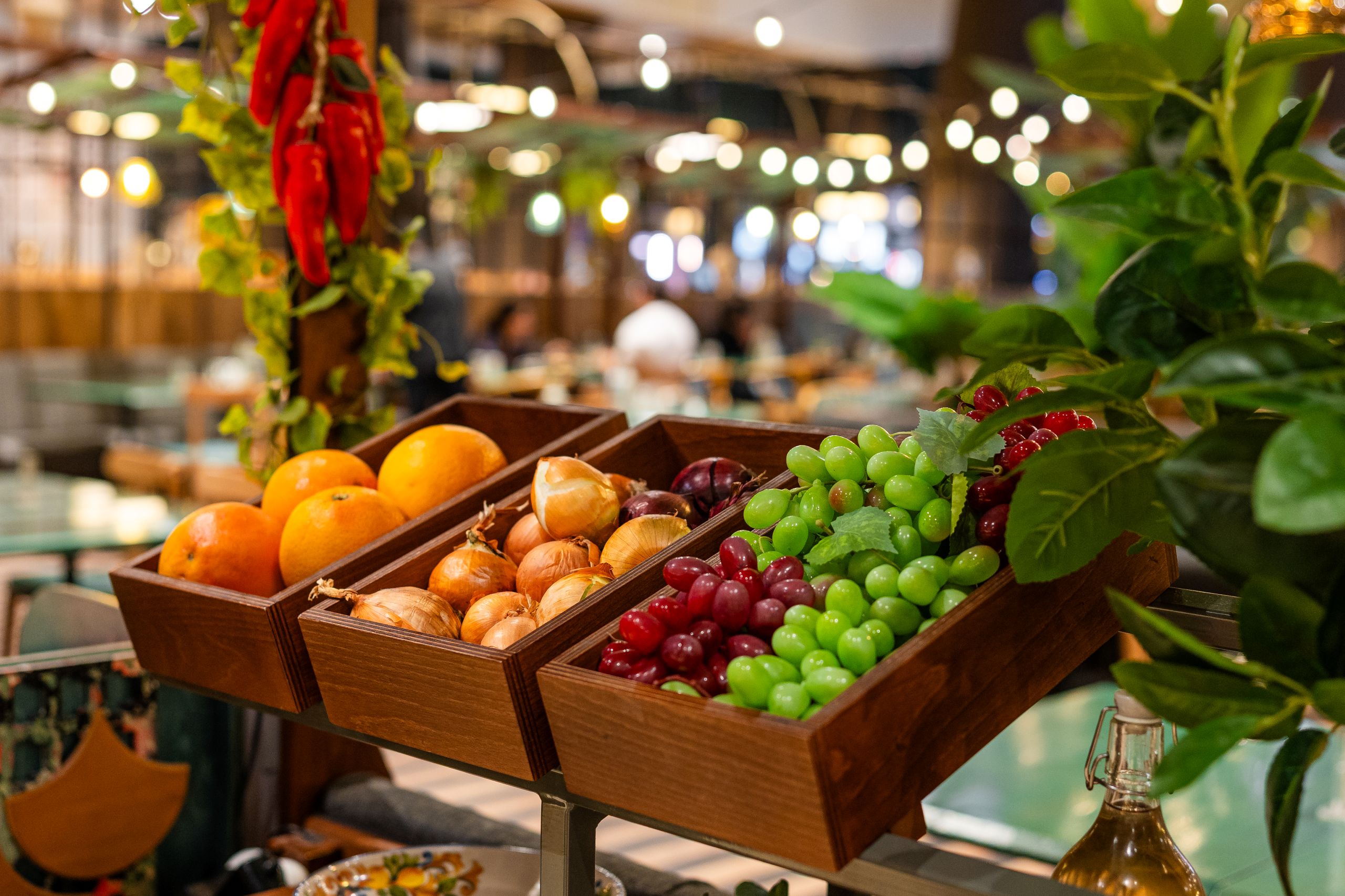 Wooden crates filled with oranges, onions, and grape clusters on a market counter with a warm, blurred background.