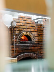 Brick pizza oven with glowing fire in the arched opening; a pizza peel rests against the brick wall.