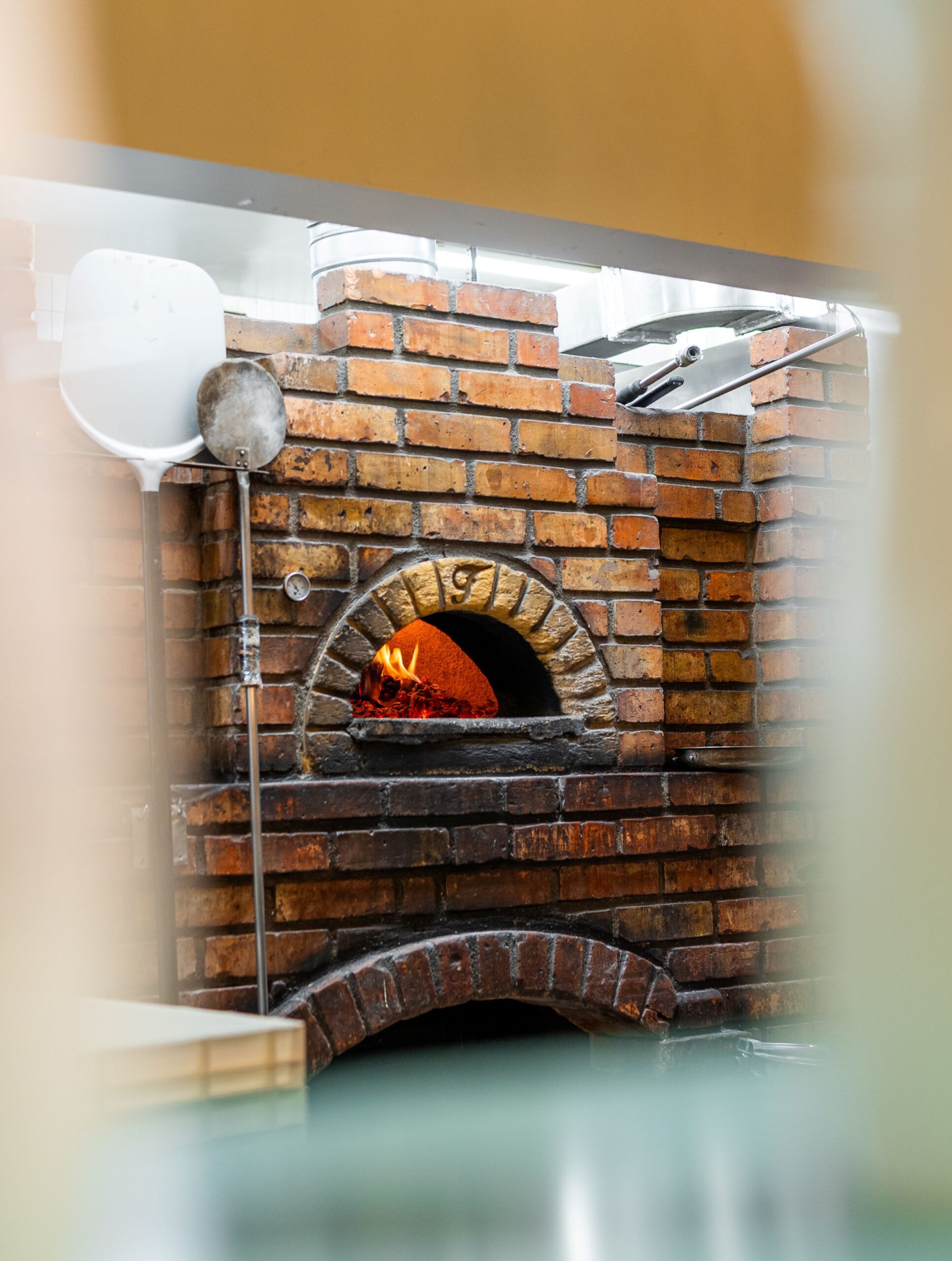 Brick pizza oven with glowing fire in the arched opening; a pizza peel rests against the brick wall.