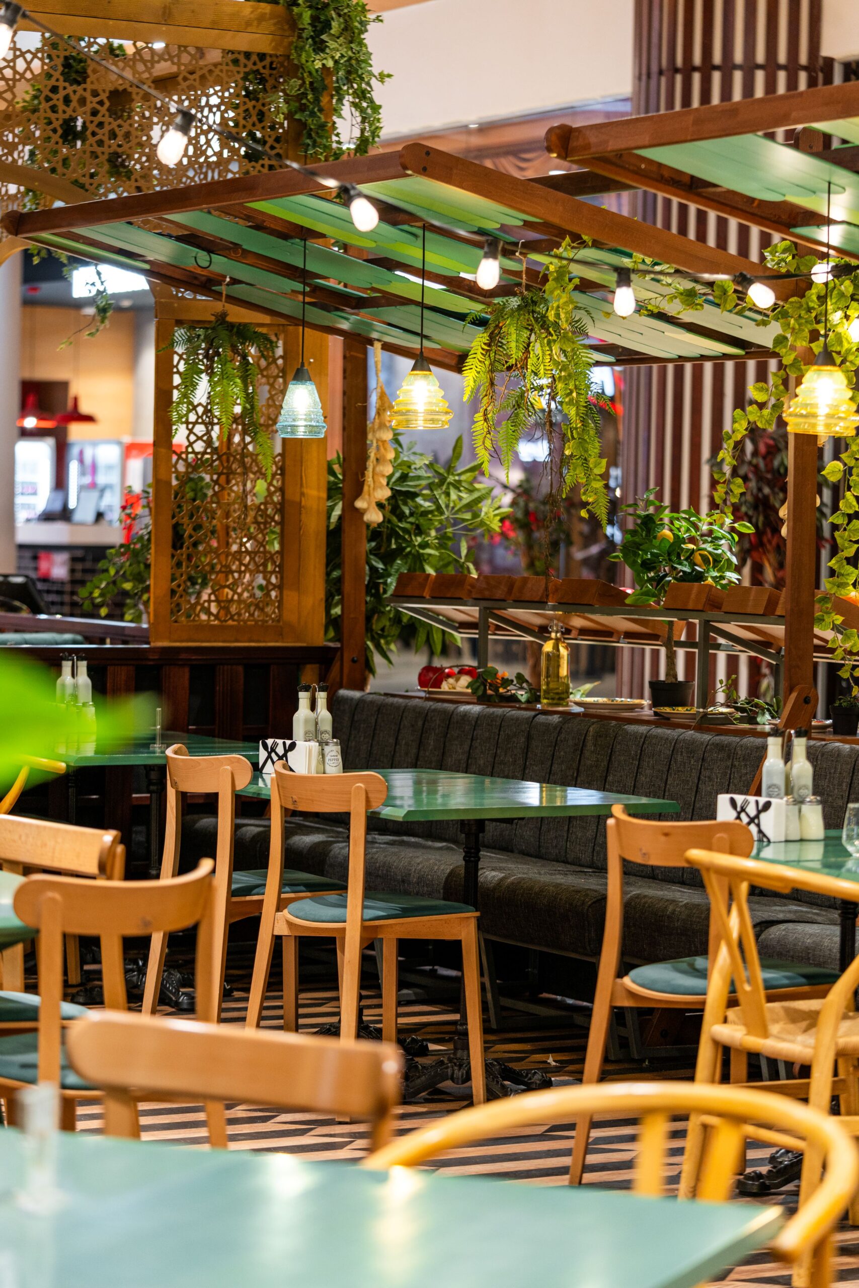 Cafe dining area with green tables, wooden chairs, hanging lights, and leafy plants in a warm, decorative space.