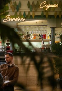 Bar interior with hanging glasses, backlit liquor shelf, and a man seated at a table beneath neon signs.