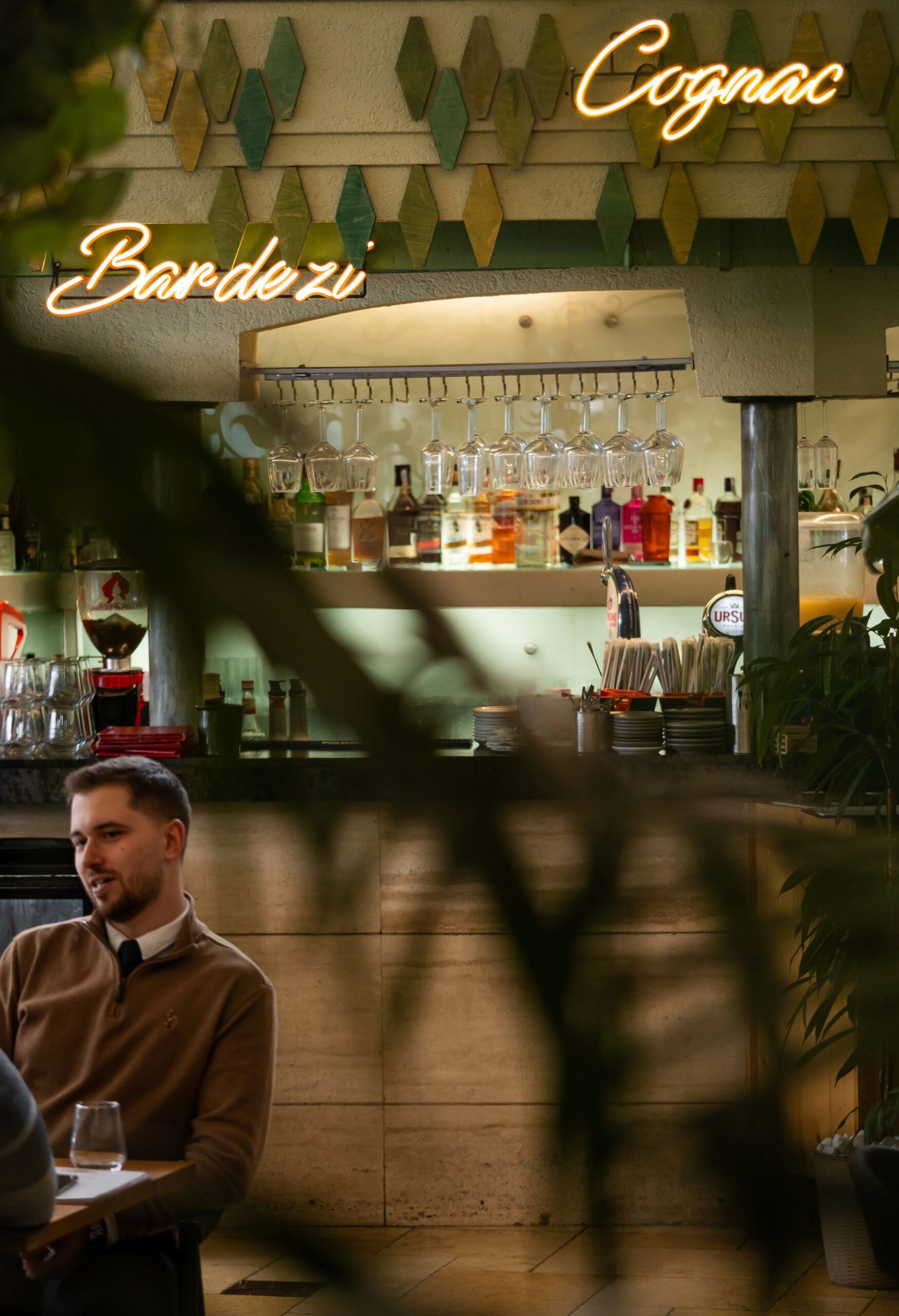 Bar interior with hanging glasses, backlit liquor shelf, and a man seated at a table beneath neon signs.