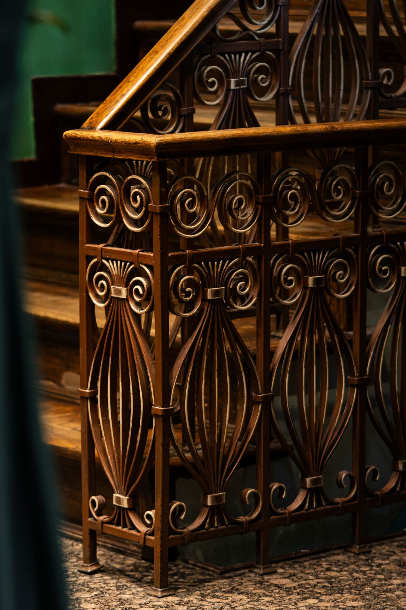 Ornate iron railing with spiral and circular designs along a wooden staircase handrail in warm lighting.