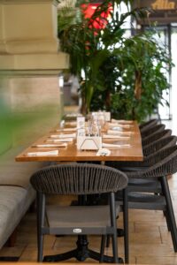 Long wooden dining table set with napkins, cutlery, and glasses, lined with wicker chairs amid lush potted plants in a bright restaurant interior.