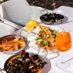 buongiorno-31 - Buongiorno Italian Outdoor table with a seafood feast: shrimp in pans, a shrimp-and-greens plate, mussels in a pan, lemon half, and an orange cocktail on a white table.
