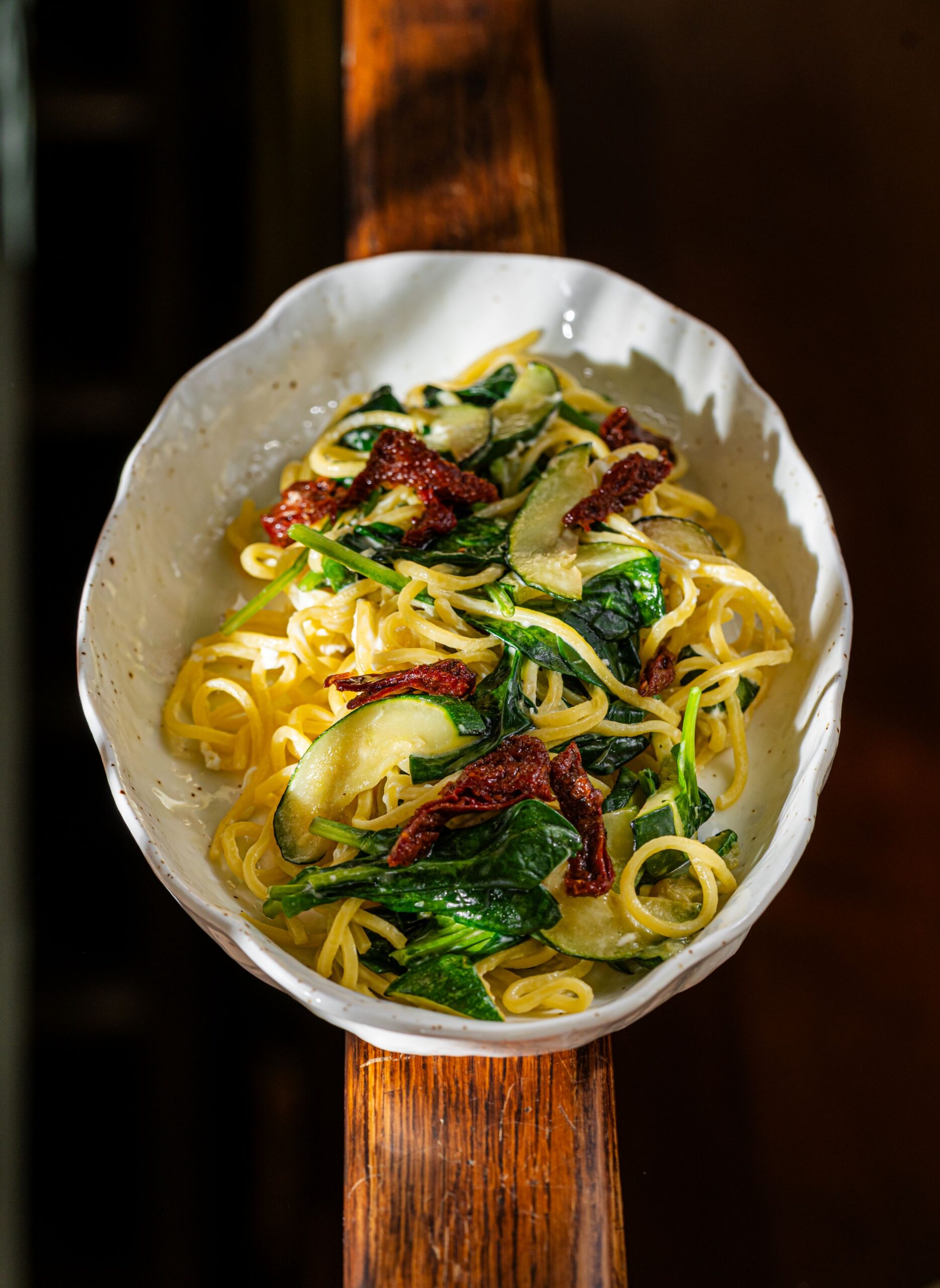 buongiorno-47 - Buongiorno Italian White ceramic bowl of pasta with spaghetti, spinach, zucchini, and sun-dried tomatoes on a wooden table.