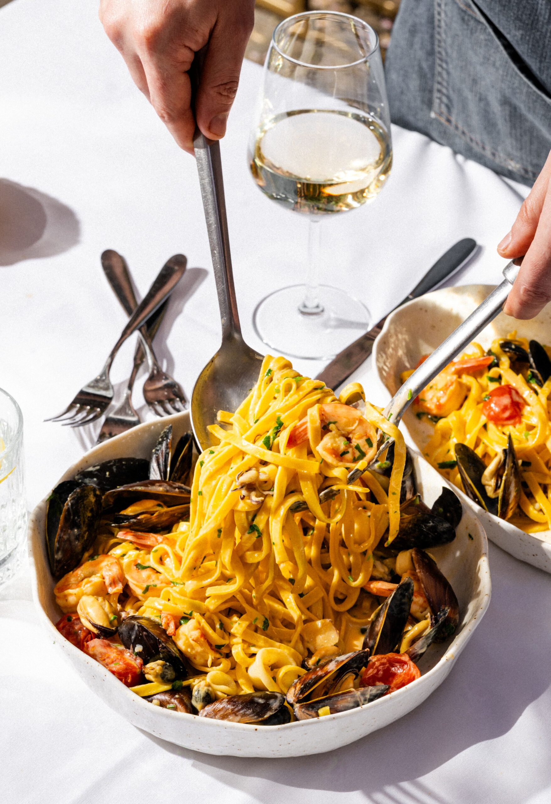 Seafood pasta with mussels and shrimp being served from a white bowl; a hand holds tongs and a glass of white wine sits nearby on the table.