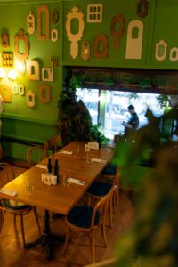 Cozy restaurant interior with lime-green walls decorated by assorted vintage frame silhouettes, wooden tables set with glassware and cutlery.