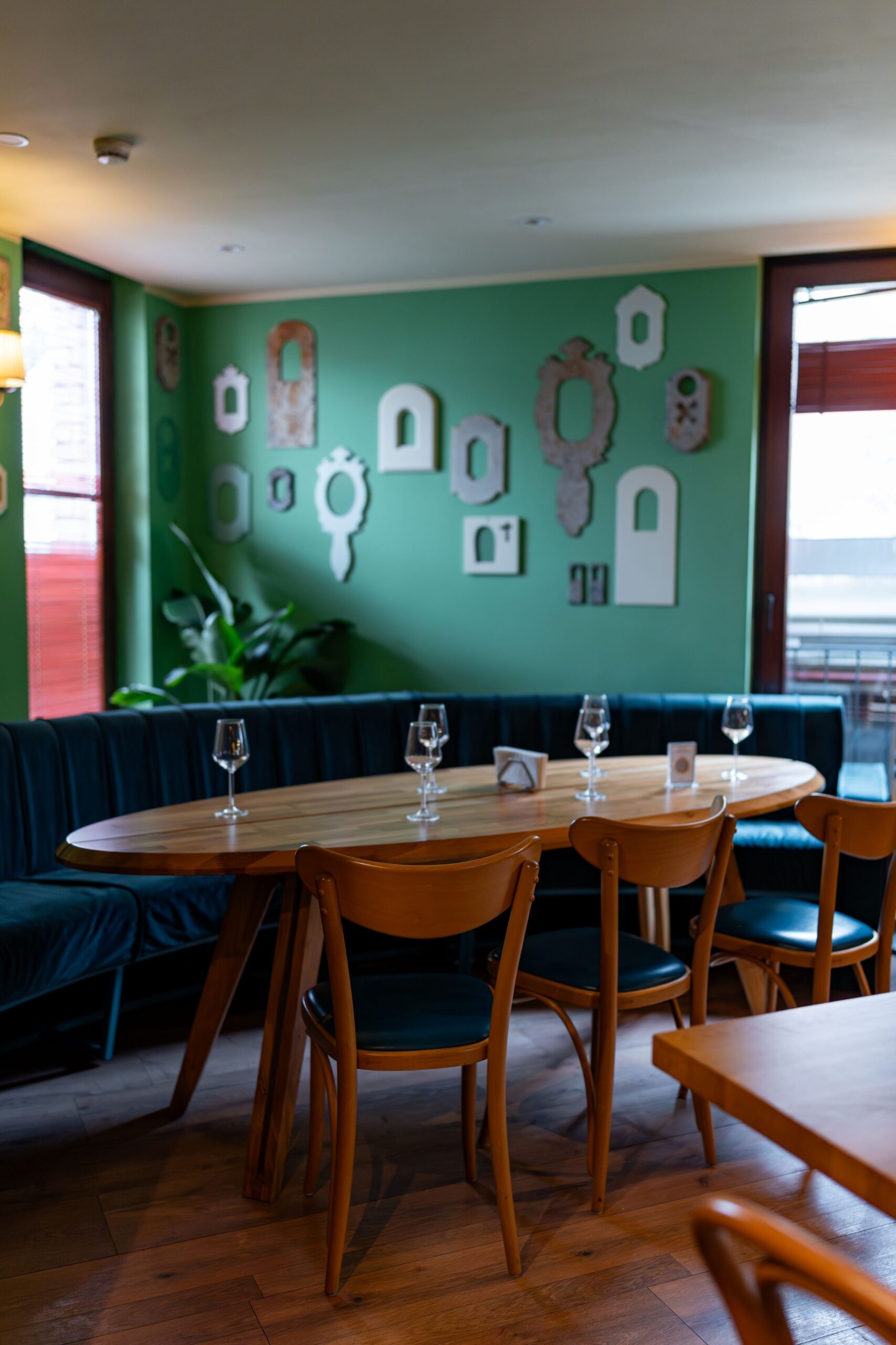photo-29 - Buongiorno Italian Dining area with a green wall decorated by various white and ornate frames, around a long wooden table set with wine glasses and chairs and a blue banquette
