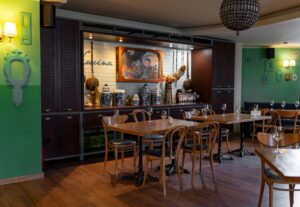 Restaurant interior with wooden tables and chairs, green walls, and a decorative bar displaying vases and glassware under warm lighting.