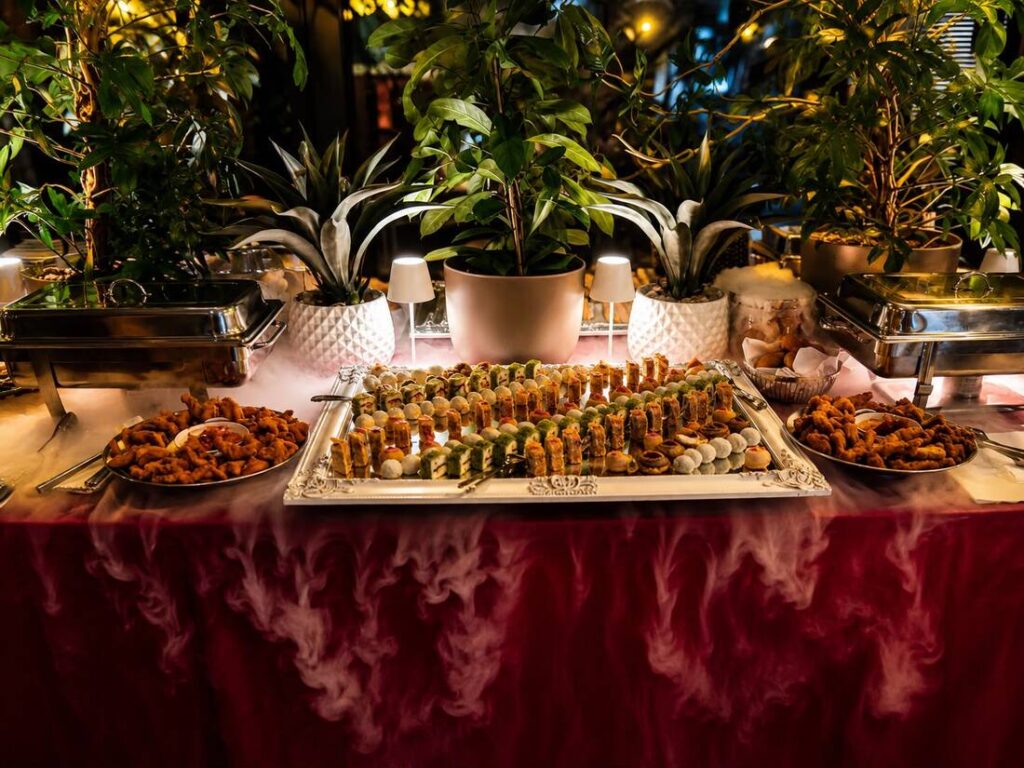 spatii private - Buongiorno Italian Buffet table with a central tray of assorted sushi rolls, surrounded by fried appetizers; potted plants and warm lighting in the background.
