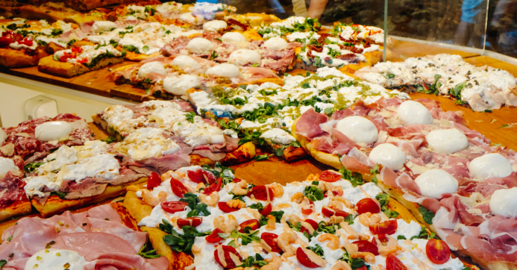 Assorted pizzas topped with mozzarella balls, ham, greens, and cherry tomatoes in a bakery display case.
