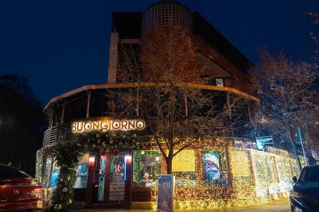 bg-primaverii1 - Buongiorno Italian Nighttime exterior of a cafe with a glowing BUONGIORNO sign and warm string lights along the storefront.
